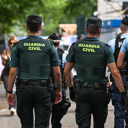 Two Guardia Civil officers, seen from behind, wear dark green uniforms with "GUARDIA CIVIL" in yellow on their backs.
