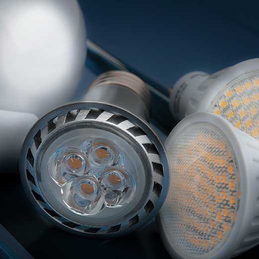 Close-up of assorted LED light bulbs on a dark blue background, highlighting a silver-rimmed spotlight with four visible LED chips in front and white bulbs with clusters of small yellow-orange diodes to the right, with the AVIENT logo in the bottom corner.