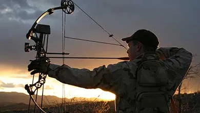 Camouflaged hunter aims compound bow in hilly sunset landscape.