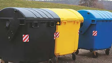 Three industrial dumpsters on wheels, black, yellow, and blue, stand outdoors.