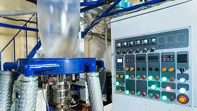 Close-up of a plastic extrusion machine showing a clear plastic tube rising beside an illuminated control panel.