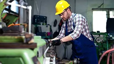 Factory worker in a yellow hard hat and blue apron operating large industrial machinery.