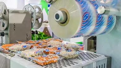 Close-up of a food packaging machine with a large roll of printed plastic film feeding in and finished vacuum-sealed food packages coming out.