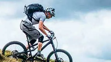 A mountain biker wearing a helmet and large backpack rides up a steep grassy hill beneath a cloudy sky.