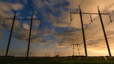 Silhouetted electrical towers and power lines stand in an open field under a twilight sky with orange and blue clouds.