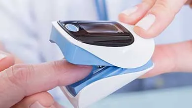 A white and blue pulse oximeter clipped on a fingertip, with a hand adjusting the device. The small digital screen displays oxygen saturation levels.