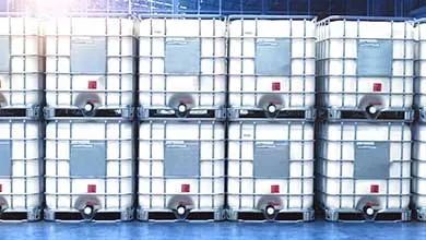 Rows of large white industrial bulk containers with metal cages, stacked two high on a blue floor, each with a rectangular label and a bottom discharge valve.